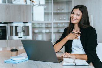 Adorable shy brunette businesswoman in black suit sitting at desk working at home using laptop